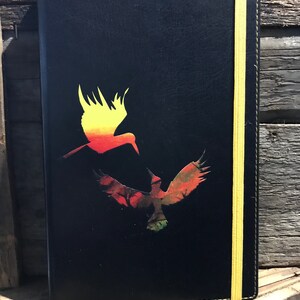 A black A5 faux soft touch leather notebook with a yellow elastic closure and yellow stitching.  The notebook has a design in the middle of two hummingbirds, one yellow and orange ombre and the other green and brown and red.