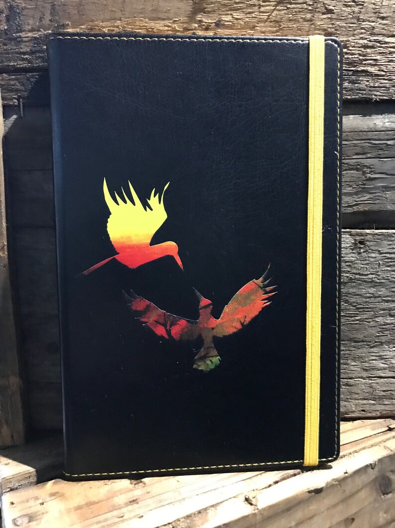 A black A5 faux soft touch leather notebook with a yellow elastic closure and yellow stitching.  The notebook has a design in the middle of two hummingbirds, one yellow and orange ombre and the other green and brown and red.