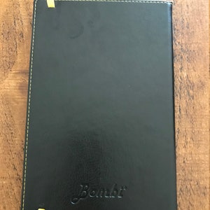 The back of the notebook is plain black but with the ends of the yellow elastic visible, the yellow stitching around the edge, and the Bombi logo is embossed in the bottom middle area of the book.