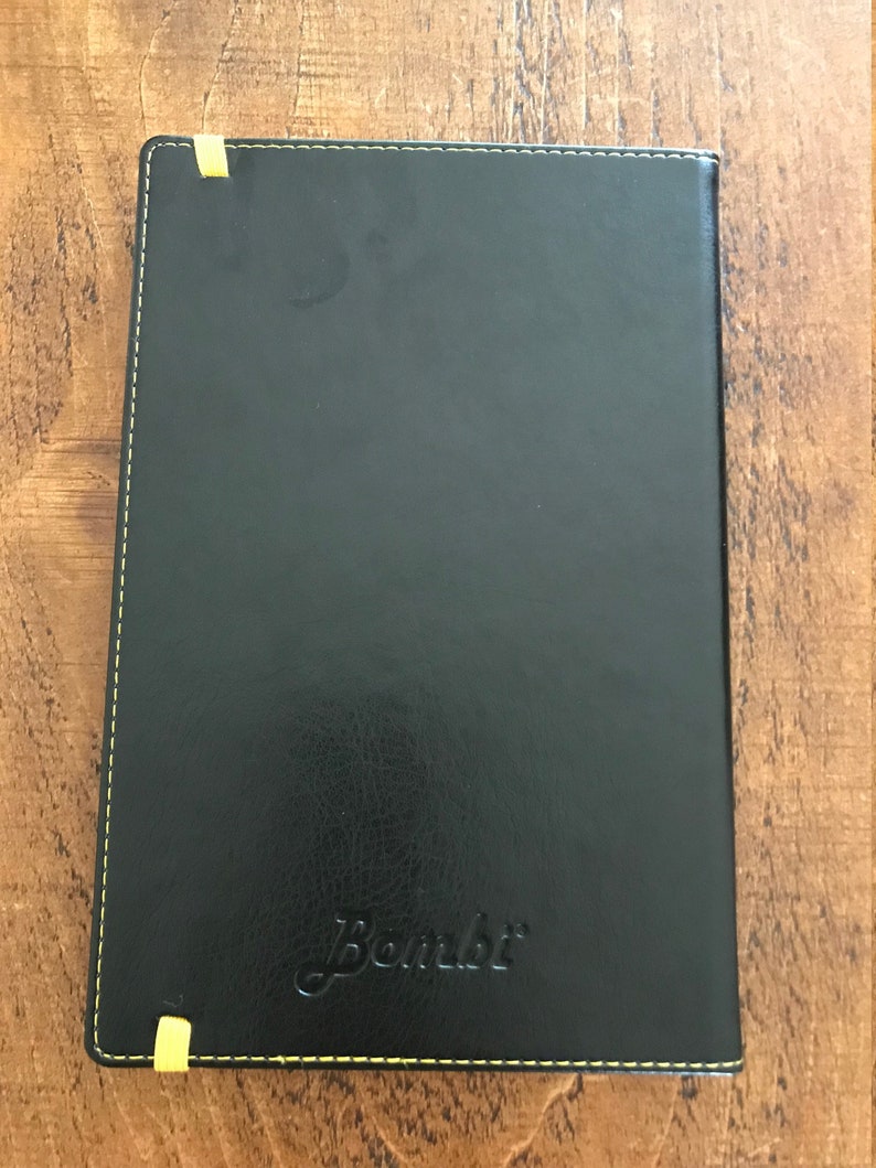 The back of the notebook is plain black but with the ends of the yellow elastic visible, the yellow stitching around the edge, and the Bombi logo is embossed in the bottom middle area of the book.