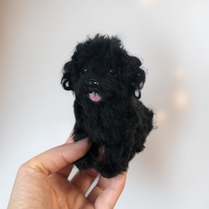 Puede incluir: Una figurita de perro de fieltro de aguja negra. El perro está sentado con la lengua fuera. La figurita está siendo sostenida en la mano de una persona.