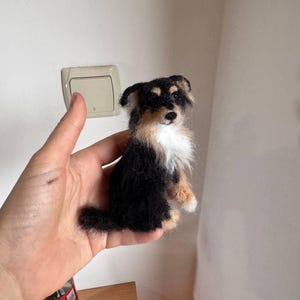 May include: A small, handmade felt dog figurine. The dog has black, tan, and white fur, with a fluffy chest and tail. The figurine is held in a hand, showcasing its size and detail. The dog's features are carefully crafted, giving it a lifelike appearance.