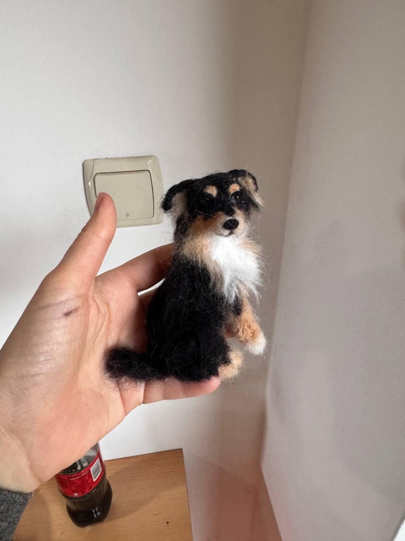 May include: A small, handmade felt dog figurine. The dog has black, tan, and white fur, with a fluffy chest and tail. The figurine is held in a hand, showcasing its size and detail. The dog's features are carefully crafted, giving it a lifelike appearance.
