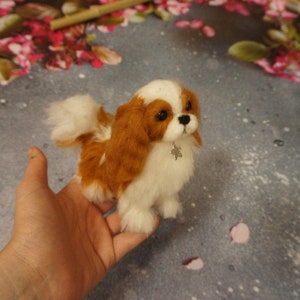 May include: A small, white and brown needle felted dog with a silver charm around its neck. The dog is sitting on a hand.