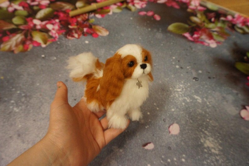 May include: A small, white and brown needle felted dog with a silver charm around its neck. The dog is sitting on a hand.