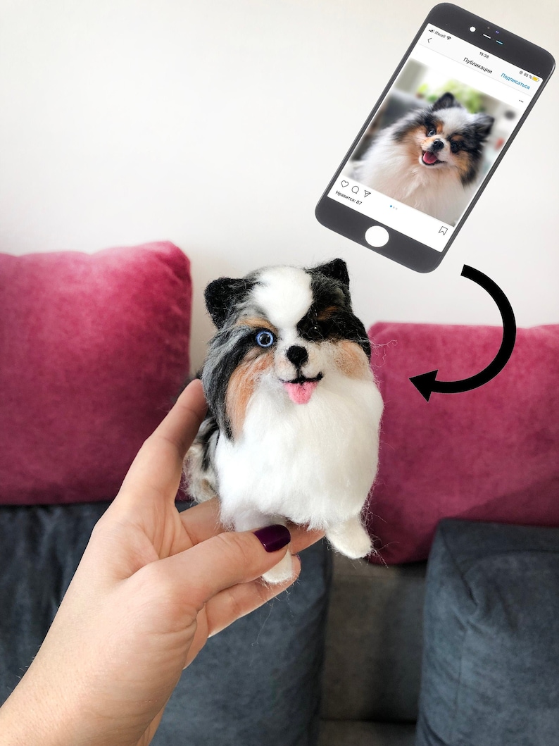 May include: A hand holds a needle felted dog toy. The toy is black, white, and brown with blue eyes. The toy is a replica of a dog in a photo on a smartphone.