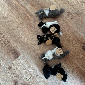May include: Five handmade cat ornaments in various colors, including gray, black, and calico, each with a small tag that says "Thank You". The cat figurines are lying down, with a string attached for hanging.