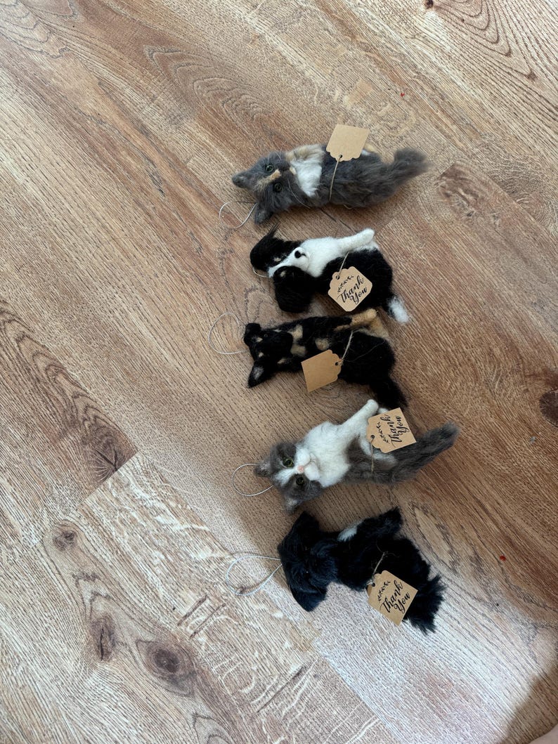 May include: Five handmade cat ornaments in various colors, including gray, black, and calico, each with a small tag that says "Thank You". The cat figurines are lying down, with a string attached for hanging.