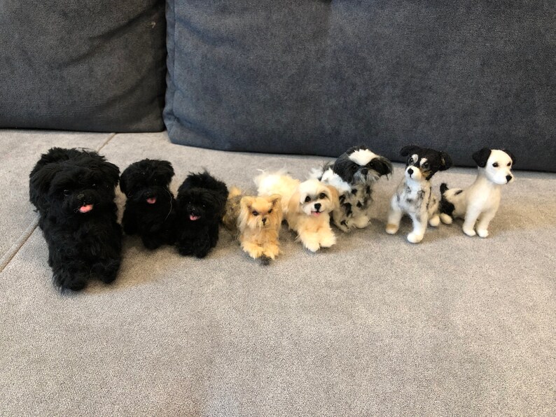 May include: A row of eight needle felted dog figurines. The figurines are all different breeds and colors, including black, white, brown, and gray. The figurines are arranged in a line on a gray couch.