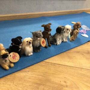 May include: A row of eight felted animal figurines, including dogs and a cat, arranged on a blue mat. Each figurine has a small tag with a heart design.