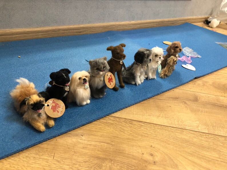 May include: A row of eight felted animal figurines, including dogs and a cat, arranged on a blue mat. Each figurine has a small tag with a heart design.