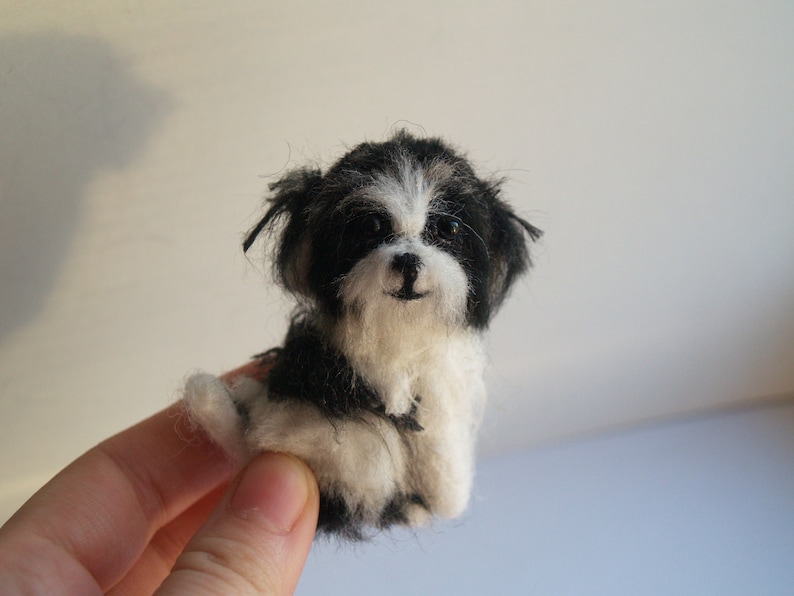 May include: A small, handmade dog figurine with a fluffy, textured appearance. The dog is primarily white with patches of black and gray fur. It has small, dark eyes and a black nose, and is sitting upright.