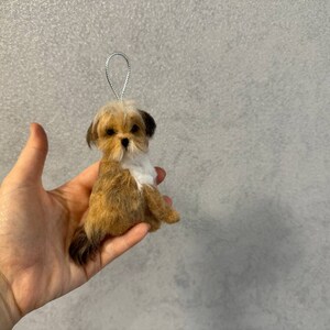 May include: A small, handmade dog ornament with a silver loop for hanging. The dog is brown and white, with a fluffy appearance. The ornament is held in a hand against a gray background.