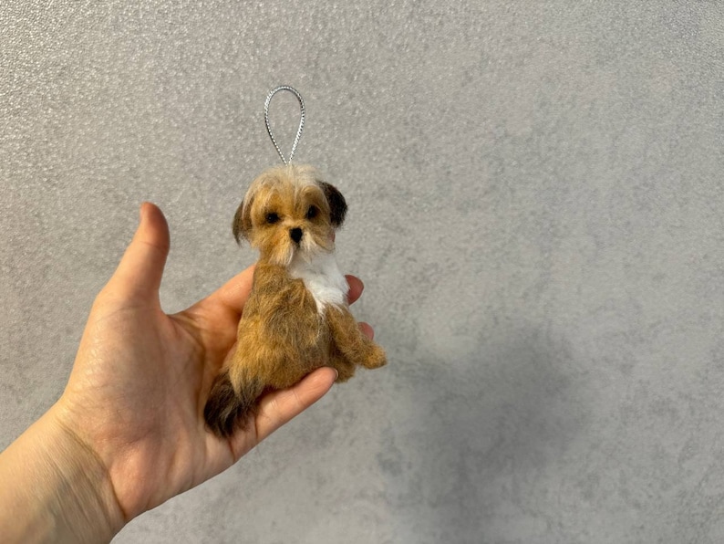 May include: A small, handmade dog ornament with a silver loop for hanging. The dog is brown and white, with a fluffy appearance. The ornament is held in a hand against a gray background.