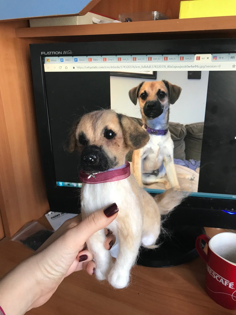 May include: Two needle felted dog figurines. The smaller dog is being held in a hand. The larger dog is sitting on a brown surface. Both dogs are brown and white with black noses and eyes. The smaller dog is wearing a burgundy collar with a small bow.