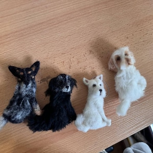 May include: Four handmade felt dog figurines in various breeds. The dogs are in shades of black, white, and brown, with detailed features. The figurines are arranged on a wooden surface, showcasing their unique designs and textures.