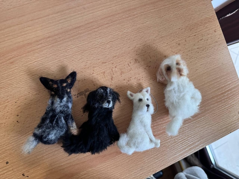 May include: Four handmade felt dog figurines in various breeds. The dogs are in shades of black, white, and brown, with detailed features. The figurines are arranged on a wooden surface, showcasing their unique designs and textures.