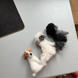 May include: Three small, handmade dog figurines. One is white with tan markings, another is white with gray markings, and the third is black and gray. The dogs are arranged in a row on a light gray surface.