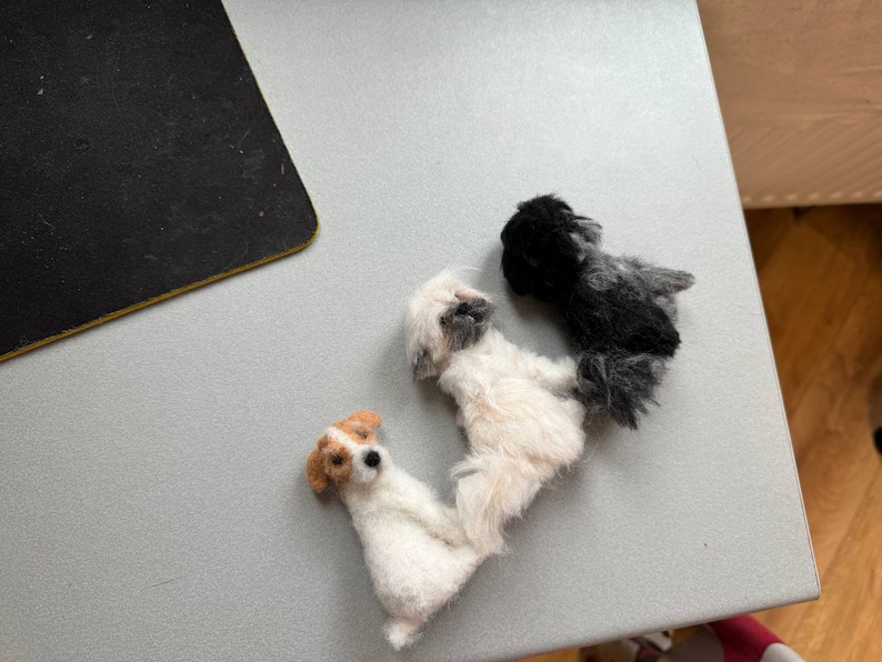 May include: Three small, handmade dog figurines. One is white with tan markings, another is white with gray markings, and the third is black and gray. The dogs are arranged in a row on a light gray surface.