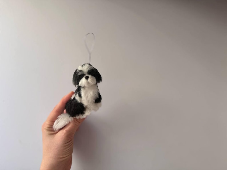 May include: A black and white Shih Tzu dog ornament with a loop for hanging. The small dog figurine is made of soft material and has a fluffy tail. The dog is sitting upright and has a sweet expression.