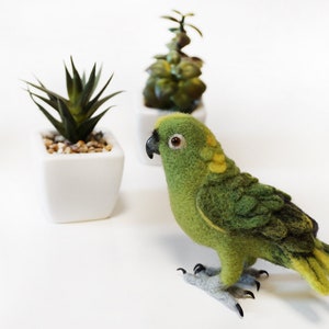 May include: A green and yellow felt parrot with a black beak and gray feet. The parrot is sitting on a white surface in front of two potted plants.