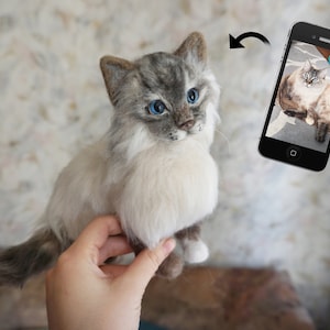 May include: A hand holds a needle felted cat with white and gray fur and blue eyes. The cat is looking at the camera. A phone is in the background showing a photo of a similar cat.