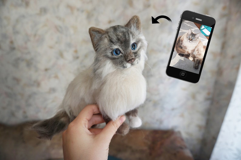 May include: A hand holds a needle felted cat with white and gray fur and blue eyes. The cat is looking at the camera. A phone is in the background showing a photo of a similar cat.