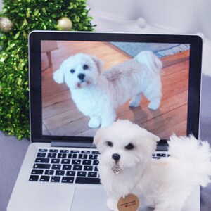 May include: A white fluffy dog toy sitting on a laptop computer with a picture of a white dog on the screen. The laptop is on a gray surface. The dog toy has a brown tag that says "Handmade with love".