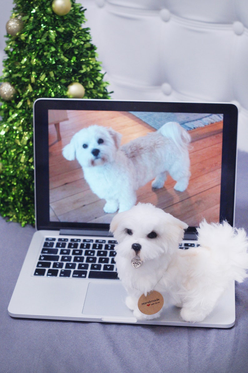 May include: A white fluffy dog toy sitting on a laptop computer with a picture of a white dog on the screen. The laptop is on a gray surface. The dog toy has a brown tag that says "Handmade with love".