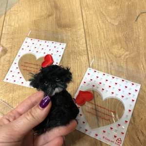 May include: A small, black plush dog with a white muzzle is held in a hand. The dog has a fluffy appearance. Two cards with heart-shaped cutouts and red heart patterns are in the background. A red heart is attached to one card.
