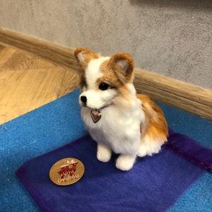 May include: A white and brown needle felted dog figurine sitting on a purple fabric surface. The dog is wearing a silver collar with a heart-shaped charm. A brown paper tag with the text "Merry Christmas" and a reindeer illustration is on the surface.