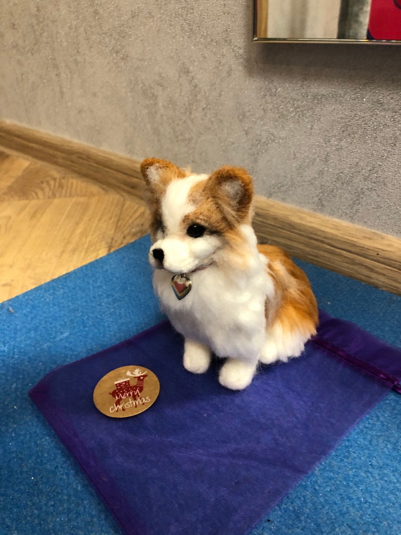 May include: A white and brown needle felted dog figurine sitting on a purple fabric surface. The dog is wearing a silver collar with a heart-shaped charm. A brown paper tag with the text "Merry Christmas" and a reindeer illustration is on the surface.