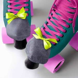 May include: Teal roller skates with pink wheels and gray toe stops. The skates have pink laces and green and pink bows.