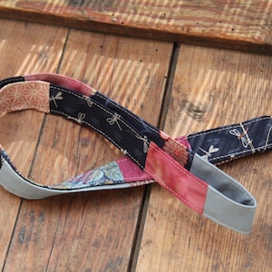 Könnte beinhalten: Eine Patchwork-Stoff-Handschlaufe mit einem silbernen Metallring. Der Riemen weist verschiedene gemusterte Stoffe in Marine, Rosa und Pfirsich auf, mit Libellen- und Blumendesigns. Die Enden sind grau.