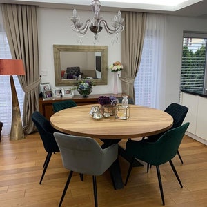 Könnte beinhalten: Ein runder Esstisch aus Holz mit einem schwarzen Metallgestell, umgeben von vier Stühlen mit dunkelgrünem und grauem Samtbezug. Der Tisch ist mit einer Blumenvase, einem Kerzenhalter und einer Dekobox gedeckt.