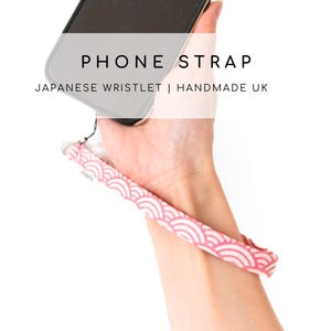 Puede incluir: Una correa de teléfono rosa y blanca con un patrón de olas japonesas. La correa está unida a una funda de teléfono negra. El texto "PHONE STRAP" y "JAPANESE WRISTLET | HANDMADE UK" es visible en la imagen.