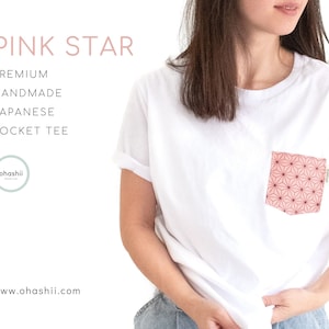以下が含まれることがあります： ピンクのポケットに幾何学的な星の模様が施された白いTシャツ。画像の上部に「PINK STAR」というテキストがあり、その下に「PREMIUM HANDMADE JAPANESE POCKET TEE」というテキストがあります。画像の左下には円の中に「ohashii」というテキストがあります。画像の下部には「www.ohashii.com」というテキストがあります。