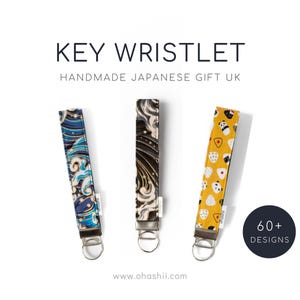 Può includere: Tre portachiavi con diversi design ispirati alla cultura giapponese. I portachiavi sono realizzati in tessuto e hanno un anello portachiavi in metallo argentato. I design includono un motivo a onde blu e bianco, un motivo a onde oro e nero e un motivo giallo con orsi panda bianchi e palline di riso. Il testo "60+ DESIGNS" si trova nell'angolo in basso a destra dell'immagine.