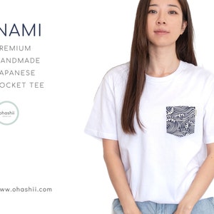 以下が含まれることがあります： 青と白の波模様のポケットが付いた白いTシャツ。 「NAMI」という文字が「PREMIUM HANDMADE JAPANESE POCKET TEE」という文字の上にあります。「ohashii」という文字が「POCKET TEE」という文字の下の円の中にあります。