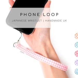 May include: A pink and white gingham phone loop attached to a black smartphone. The image includes the text "PHONE LOOP" and "JAPANESE WRISTLET | HANDMADE UK". The loop is held by a person's hand.