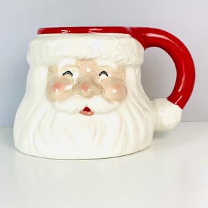 May include: A ceramic mug shaped like the face of Santa Claus, featuring a white beard, mustache, and hat trim. The mug has a glossy finish with pink cheeks, a small red mouth, and a bright red handle that extends from the top of the hat.