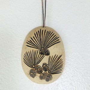 May include: An oval-shaped beige ceramic ornament featuring a dark brown etched design of pine needles and pinecones. The piece is suspended by a thin dark brown cord attached to the top, creating a decorative hanging element for display.