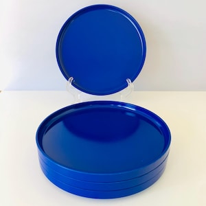 May include: A stack of four cobalt blue round plates, with a fifth plate displayed on a clear stand. The plates have a glossy finish and a slightly raised edge. The plates are a solid, vibrant blue color.
