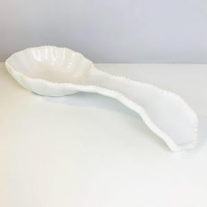 May include: A glossy white ceramic spoon rest featuring a wide, rounded bowl and an elongated handle. The edges of the spoon rest are decorated with a raised, beaded texture that follows the entire perimeter of the piece, creating a uniform look.