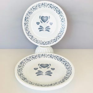 May include: Two white ceramic plates featuring a blue speckled rim pattern and a central blue graphic design consisting of one large heart flanked by two smaller hearts above four stylized leaf-like shapes arranged in a symmetrical pattern.