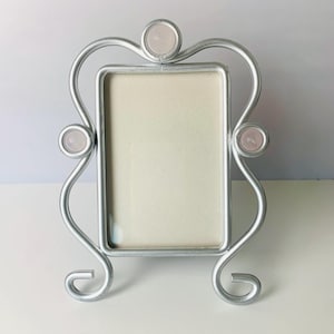 May include: Silver-toned metal picture frame with a rectangular opening. The frame has a decorative, curved design with three pink, circular accents. The frame stands on two curved feet.