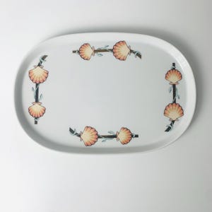 May include: White oval serving platter with a seashell design. The platter features a border of peach-colored seashells and blue-green vines. The seashells are arranged in vertical and horizontal patterns around the edge of the platter.