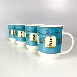 May include: Four ceramic mugs with a nautical theme. Each mug features a light blue background with a white lighthouse illustration, red anchors, and rope detailing. The mugs have white handles and bases.