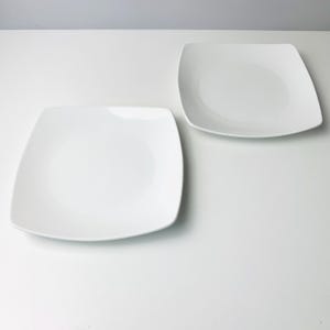 May include: Two white square plates with rounded corners are displayed on a white surface. The plates have a simple, clean design, suitable for various dining occasions. The plates are made of a glossy material.