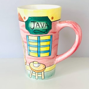 May include: A tall ceramic mug featuring a hand-painted scene of a cafe exterior. The design includes a green sign with the word JAVA, a blue-shuttered window, and a small table with a cup. The mug has a pink base, yellow top, and a pink handle.